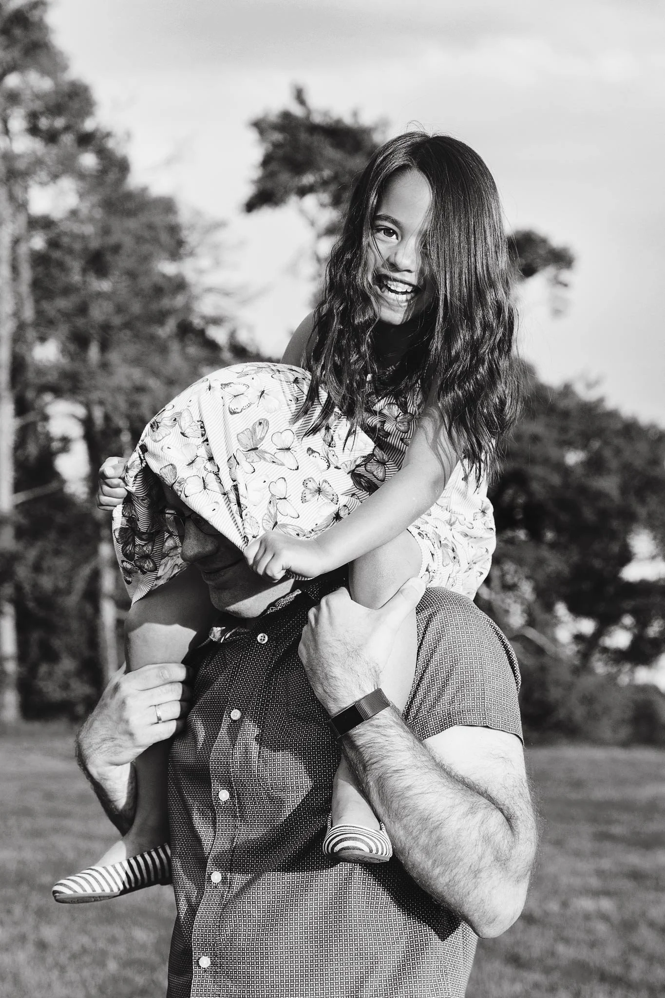 A black and white photo of a man holding a young girl on his shoulders outside in a park, smiling and laughing, with trees in the background.