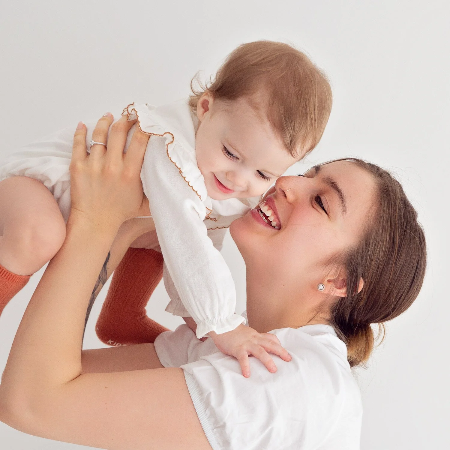 A young mother with brown hair in a bun lifts her baby girl into the air, both smiling joyfully.