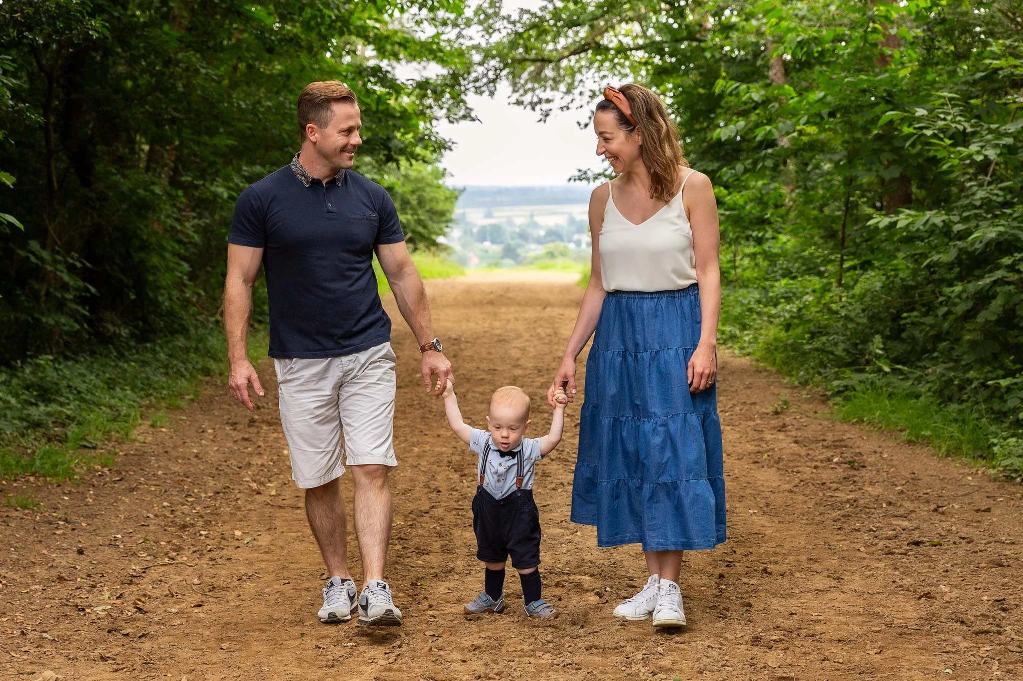 A family of three walking on a dirt path in a lush green forest. The father and mother hold the child's hands, and the child is in between them, looking forward. The father wears a navy polo shirt and shorts, the mother wears a white top and a blue s