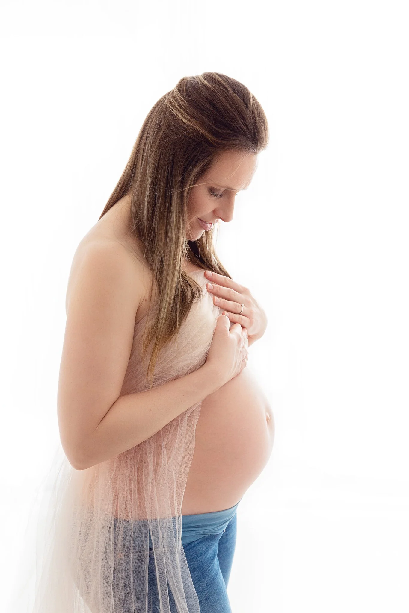 Pregnant woman with long brown hair, standing sideways, holding her belly with hands, wearing a light sheer fabric around her shoulders, against a white background.