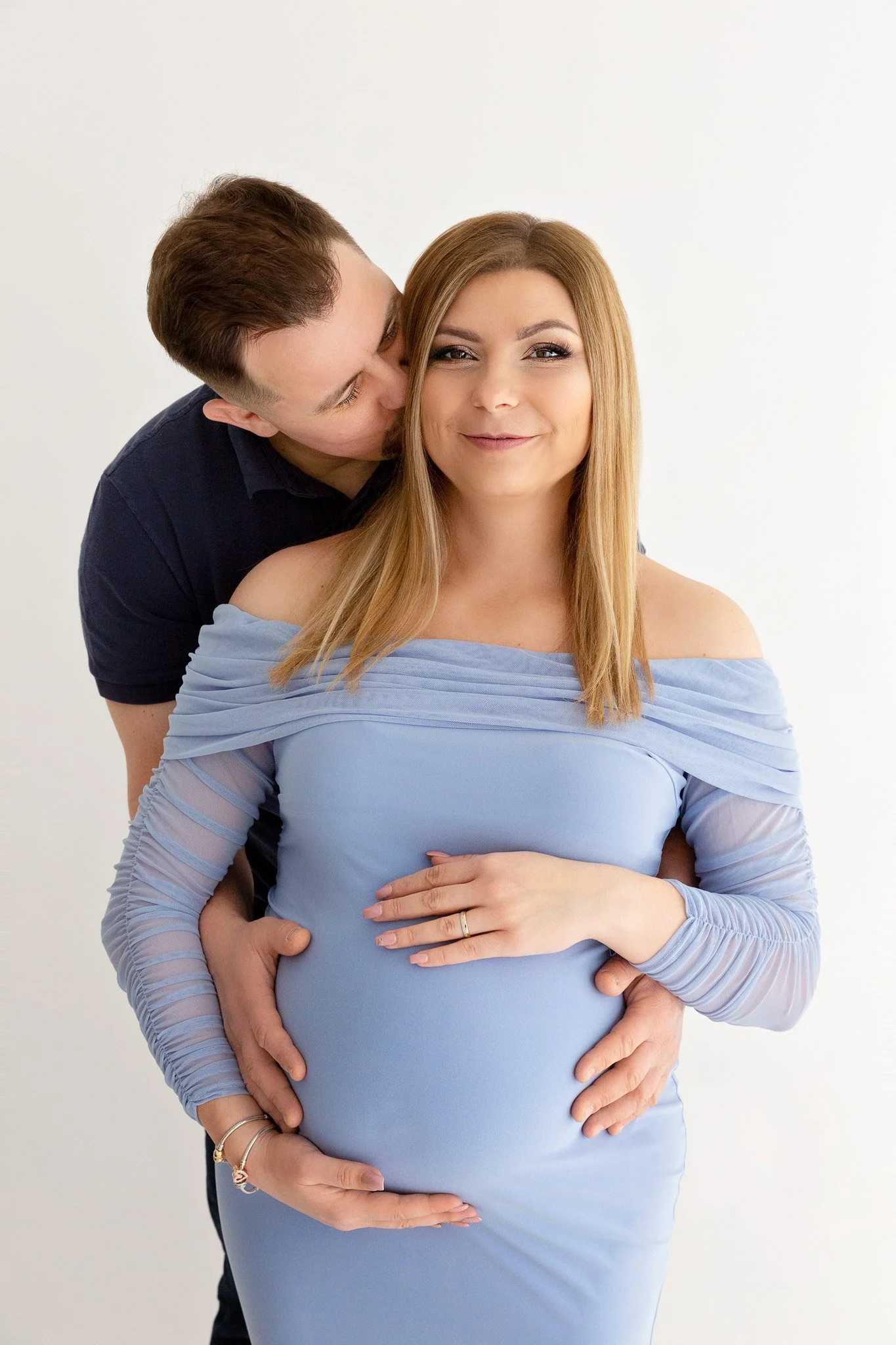 A pregnant woman in a light blue dress with an off-the-shoulder neckline stands smiling, with her partner behind her. The man, wearing a dark shirt, kisses her cheek while holding her baby bump with both hands.