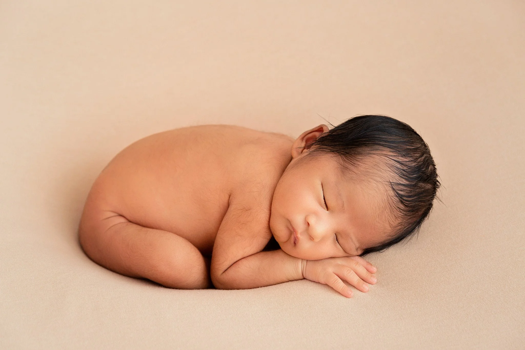 A sleeping newborn baby with dark hair laying on a beige blanket, curled into a fetal position with hands close to the face.