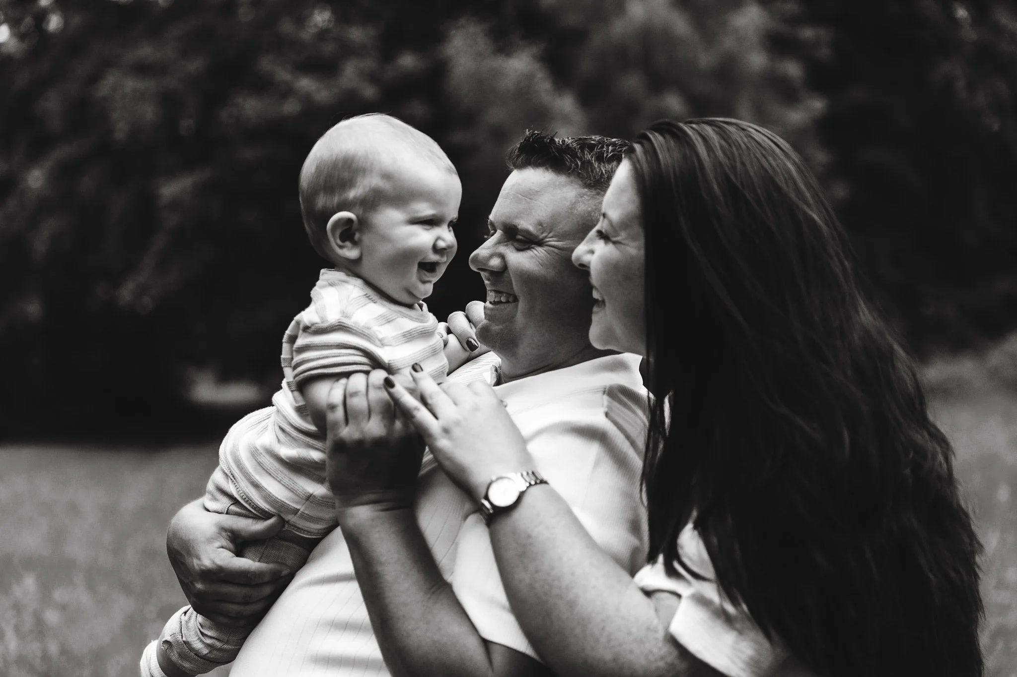 A black and white photo of two adults, a man and a woman, holding a young child between them, all smiling and laughing outdoors.