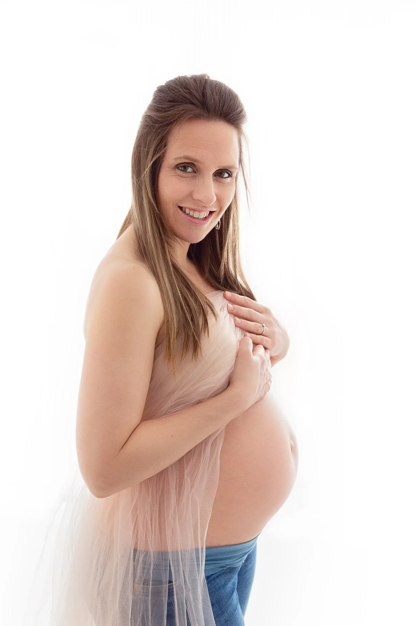 Pregnant woman smiling, partially draped in sheer fabric, holding her belly, wearing blue jeans.