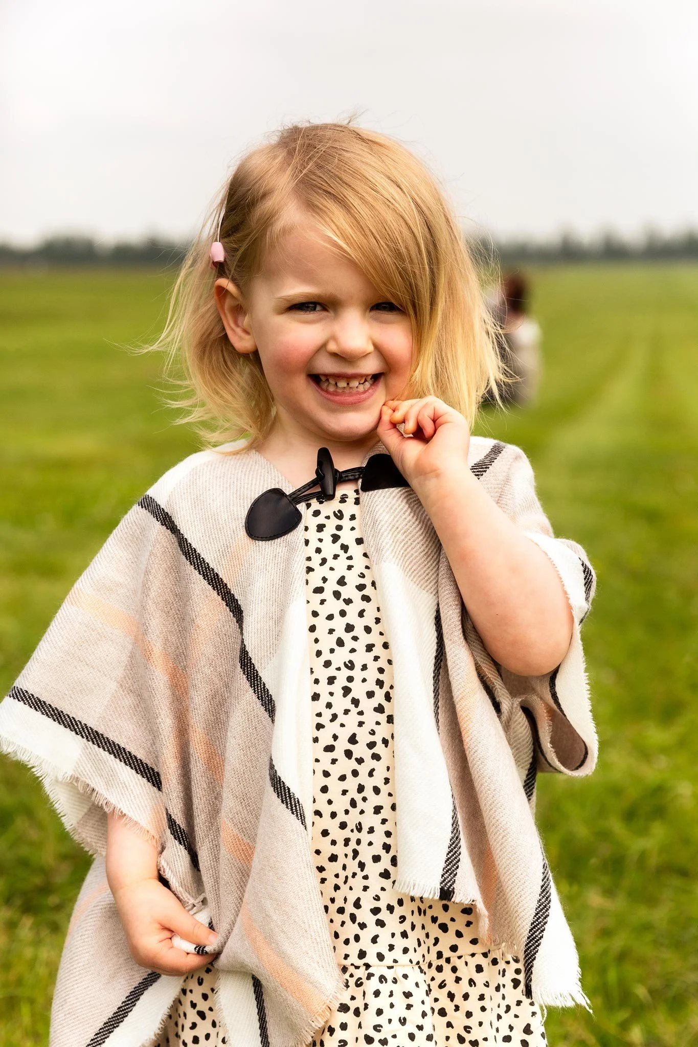 A young girl with blonde hair and a pink hair clip smiling outdoors in a grassy field, wearing a patterned dress and a beige checkered shawl.