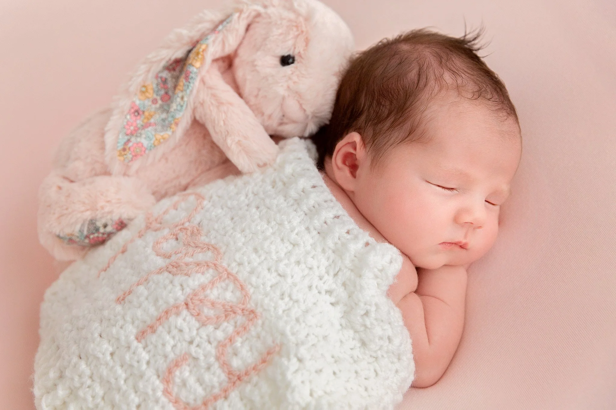 A sleeping baby with short dark hair, resting on a pink blanket, cuddled under a white knitted cardigan with pink embroidery, next to a pink plush rabbit toy with floral-patterned floppy ears.