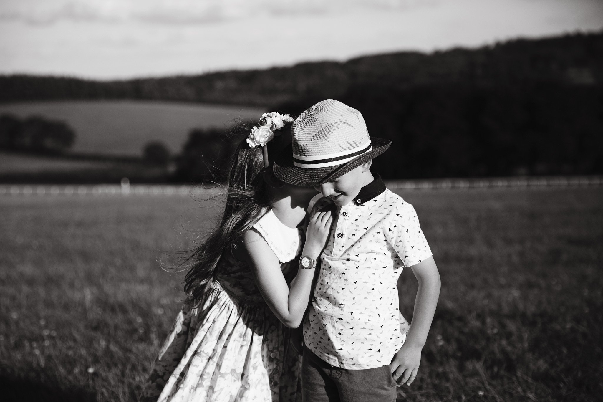 A girl and boy standing close on a grassy field, with the girl whispering into the boy's ear, both wearing summer clothes and hats, with a landscape of trees and hills in the background.