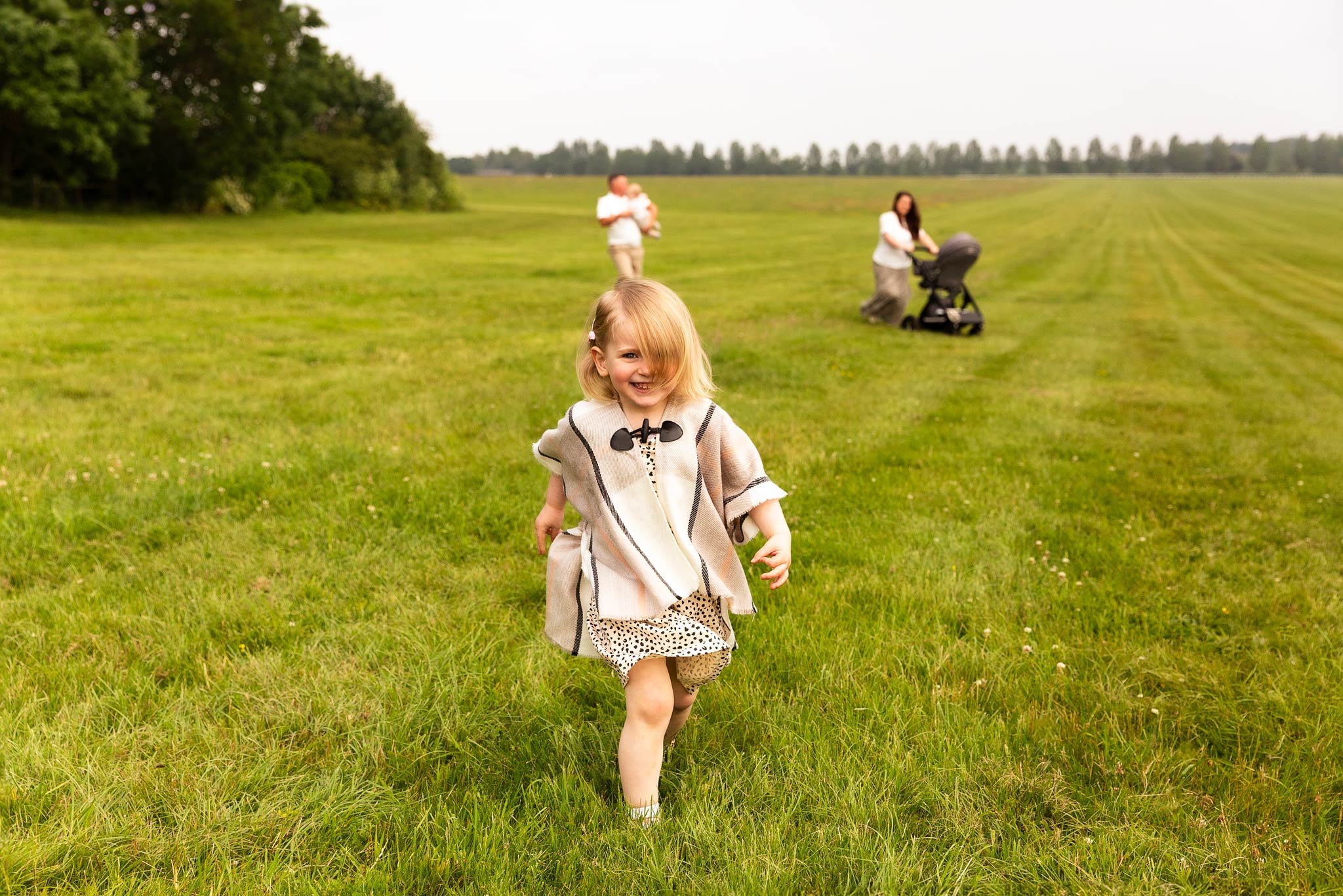 A young girl running on a grassy field, smiling, with two adults in the background walking and pushing a stroller, overlooking a wide-open meadow.