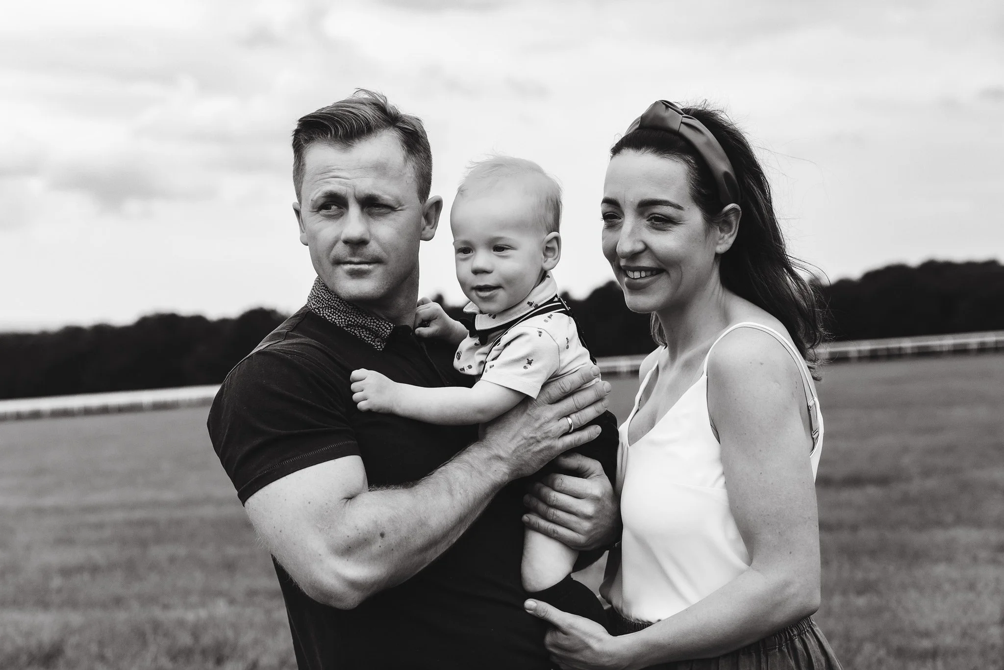 A black and white photo of a family of three outdoors. The man is holding a young child, and the woman is standing beside them, all smiling.