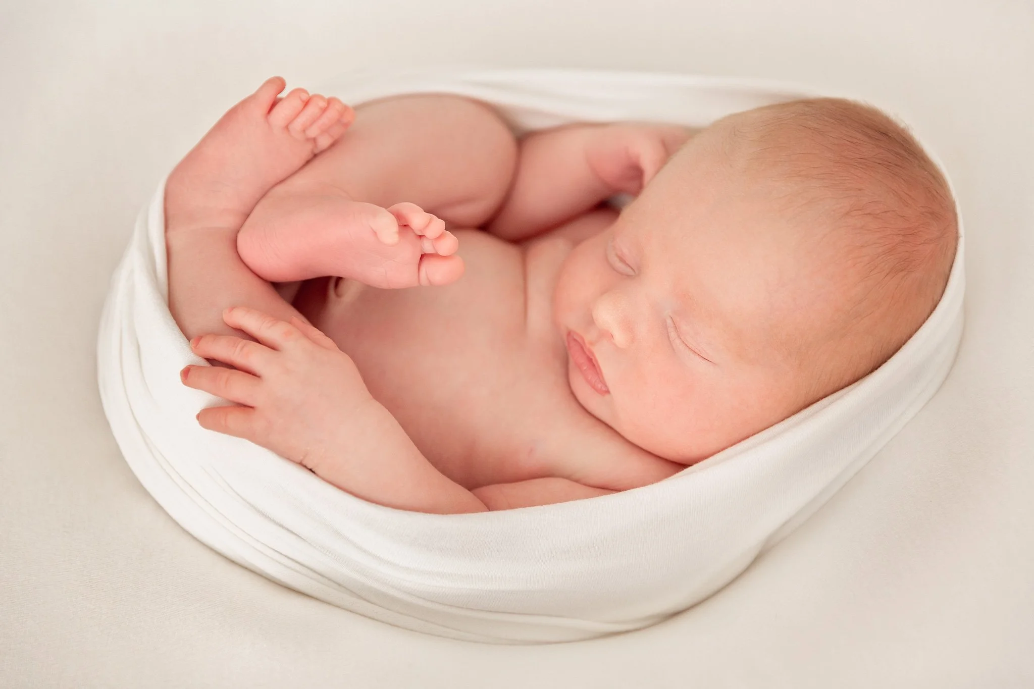 A sleeping newborn baby curled up on a soft white blanket.
