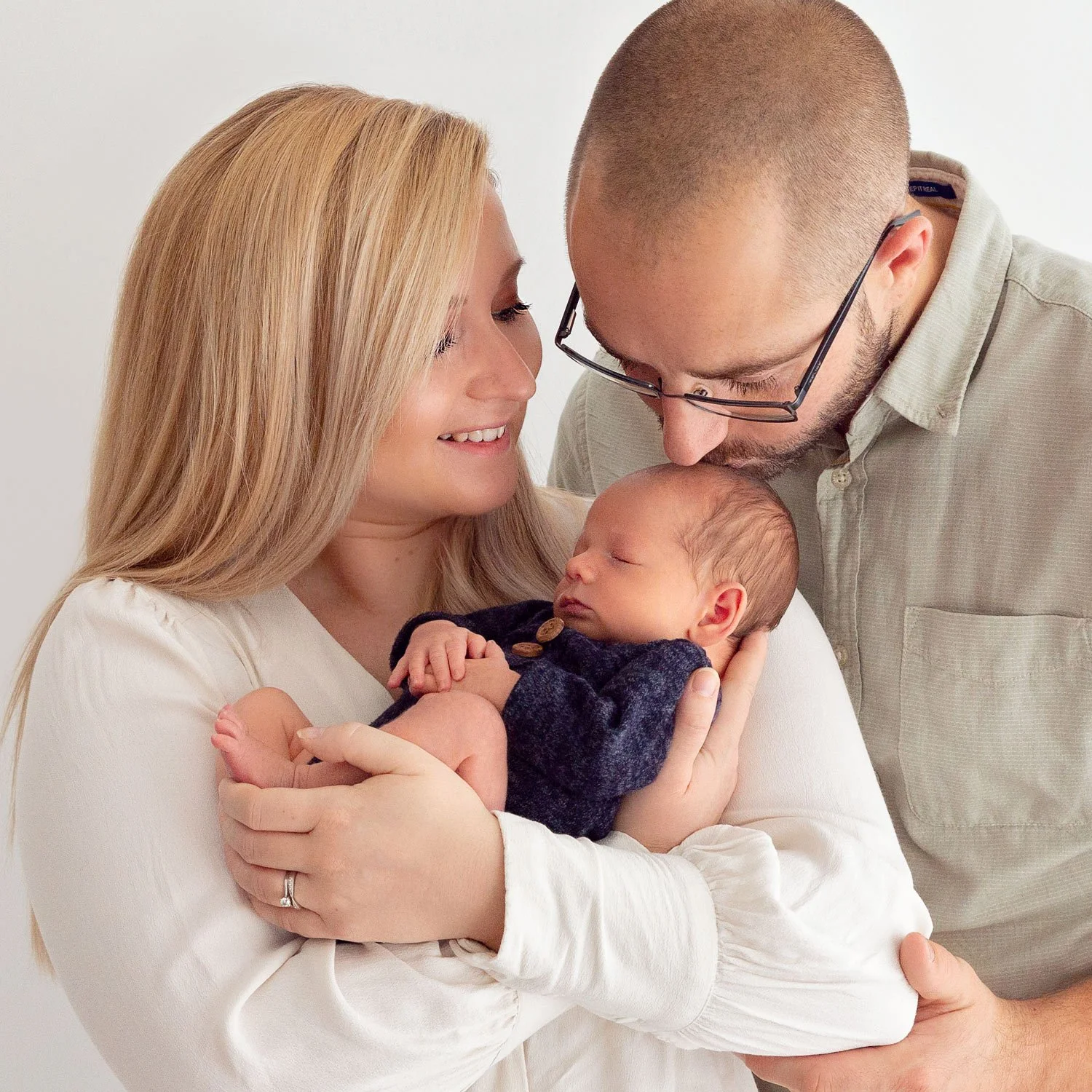 A mother with long blonde hair holds her baby as the father wraps his arms around them and kisses the baby’s head.