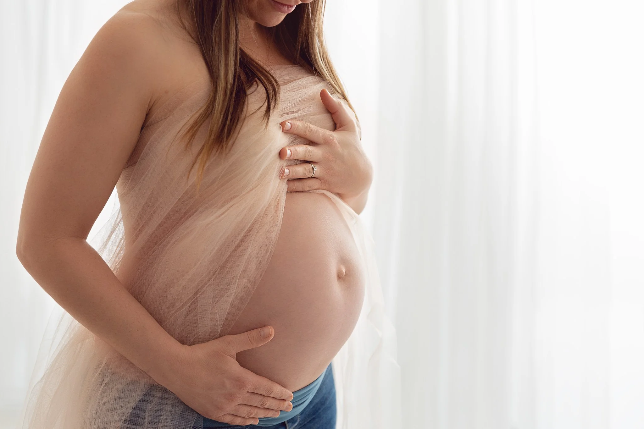 Pregnant woman with long brown hair, wearing a sheer, peach-colored fabric, gently touching her belly and chest, standing in front of a light-colored curtain background.