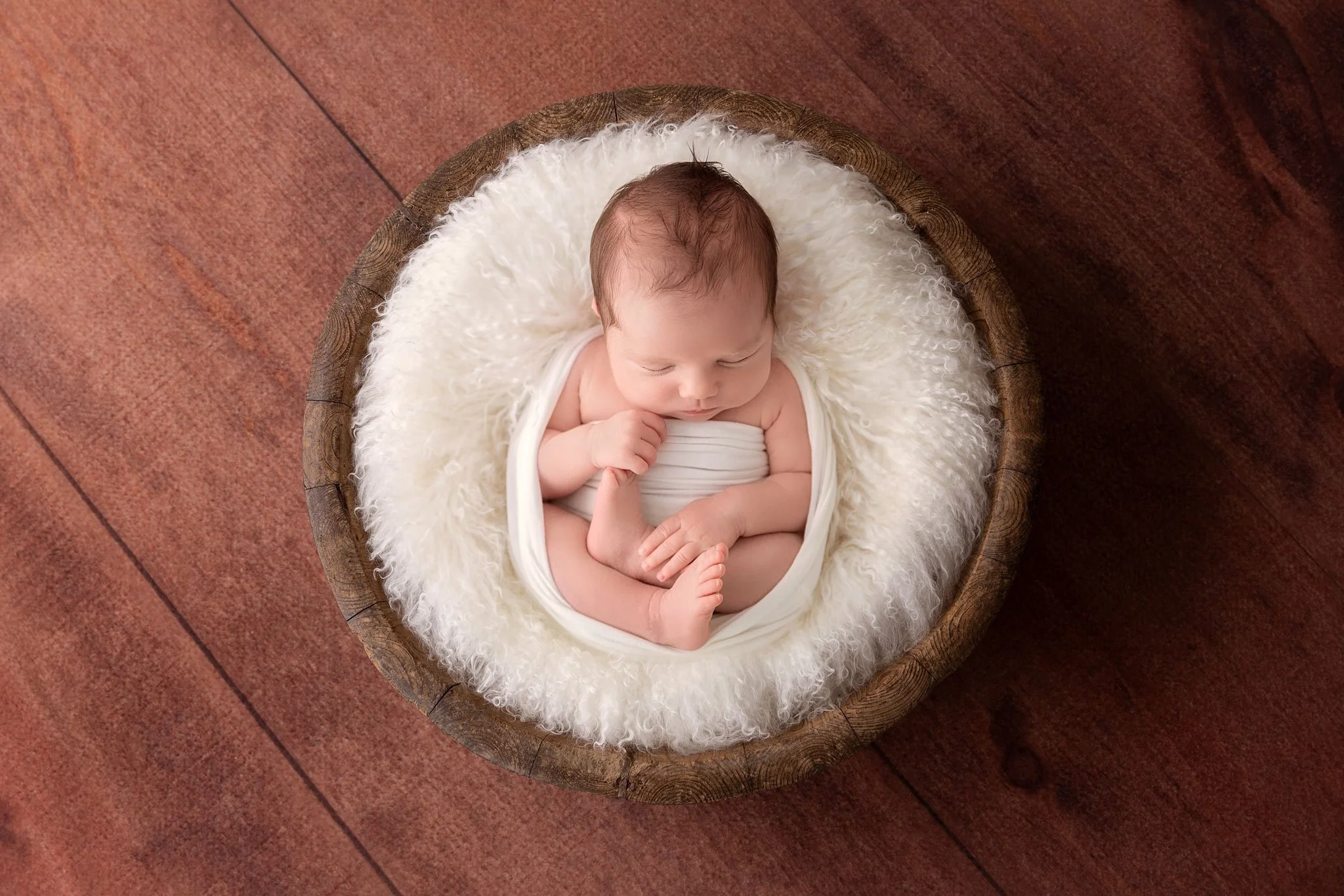 A newborn baby sleeping curled up in a white blanket inside a round wooden basket with a soft white fur lining, placed on a wooden floor.