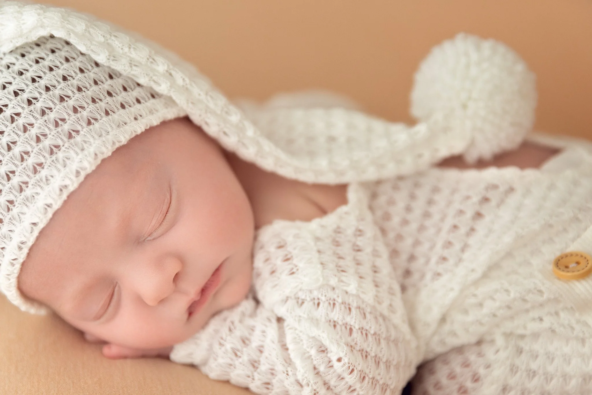 Close-up of a sleeping baby wearing a knitted white hat with a pom-pom and a matching knitted outfit.