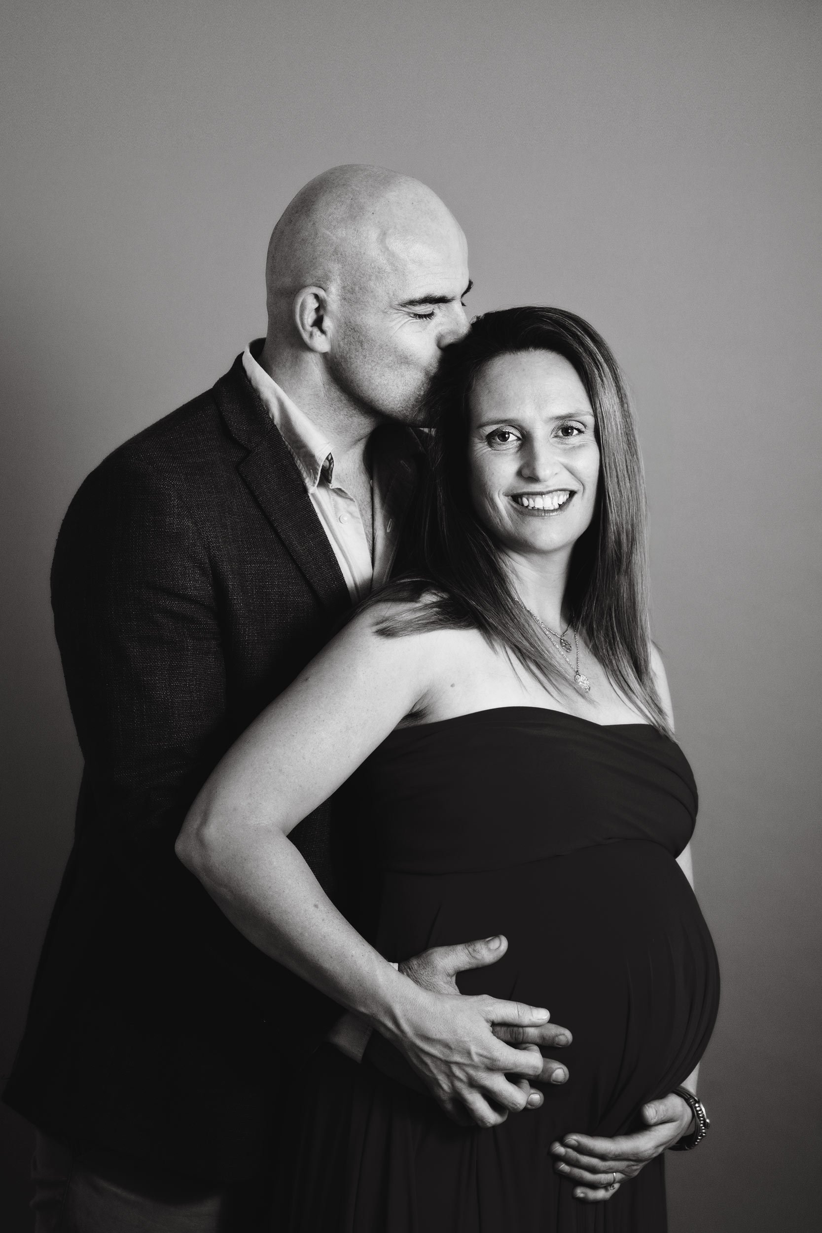 A black and white photo of a couple, with the man kissing the woman's forehead. The woman is visibly pregnant and smiling, wearing a strapless dress and jewelry. The man is wearing a suit jacket and shirt.