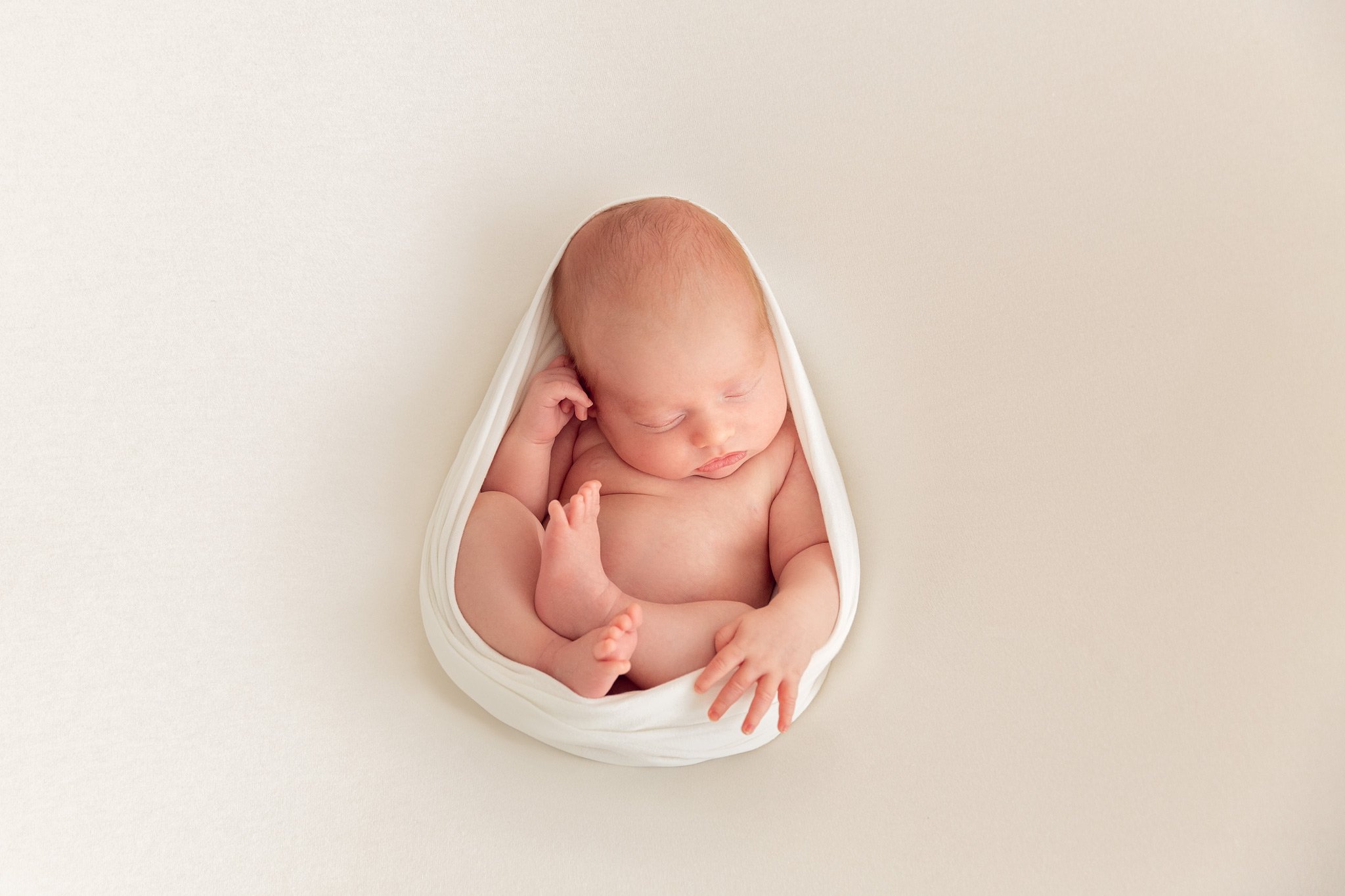 A sleeping newborn baby wrapped in a white stretchy wrap, lying on a beige blanket.