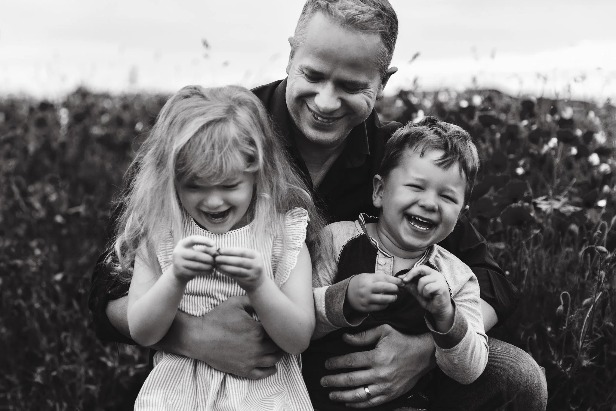 A man hugging two laughing children, a girl and a boy, outdoors in a field.