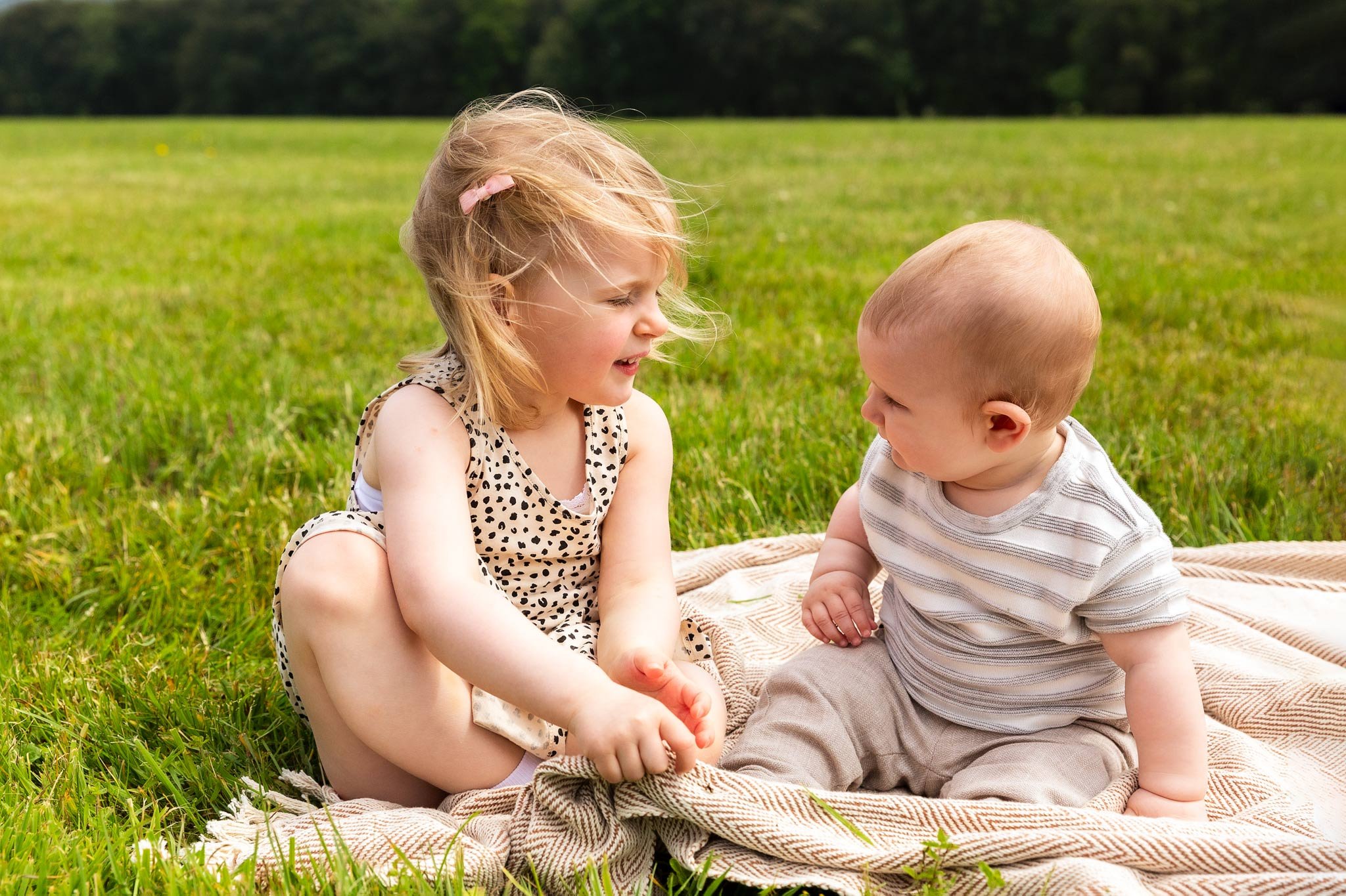 A young girl and baby sitting on a blanket in a grassy field, playing and smiling at each other on a sunny day.