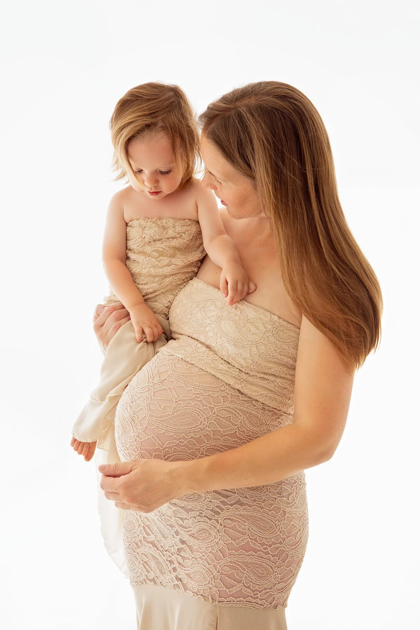 A pregnant woman holding a little girl, both wearing matching beige lace dresses, against a white background.