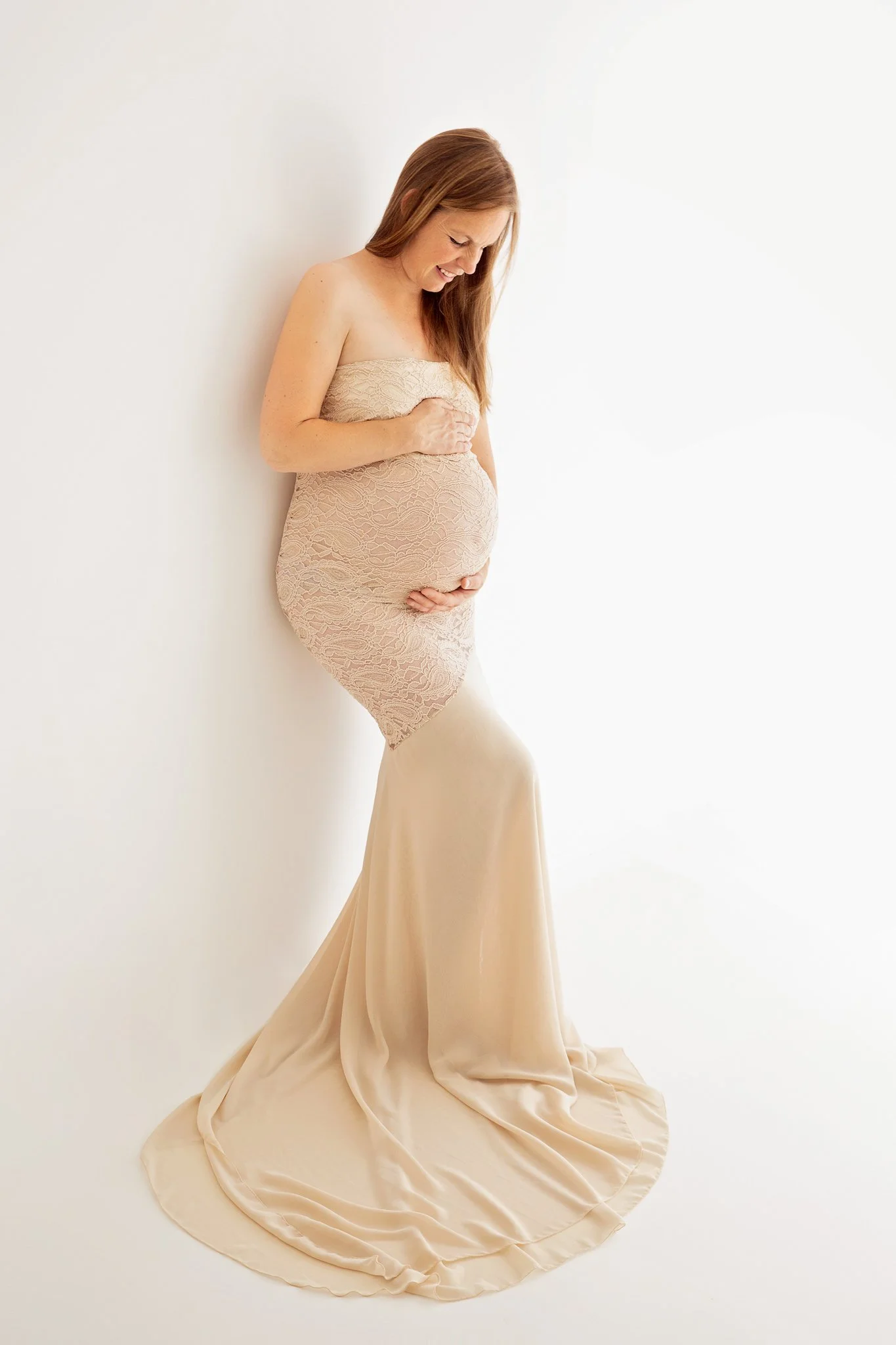 Pregnant woman wearing a beige lace dress, holding her belly and smiling.