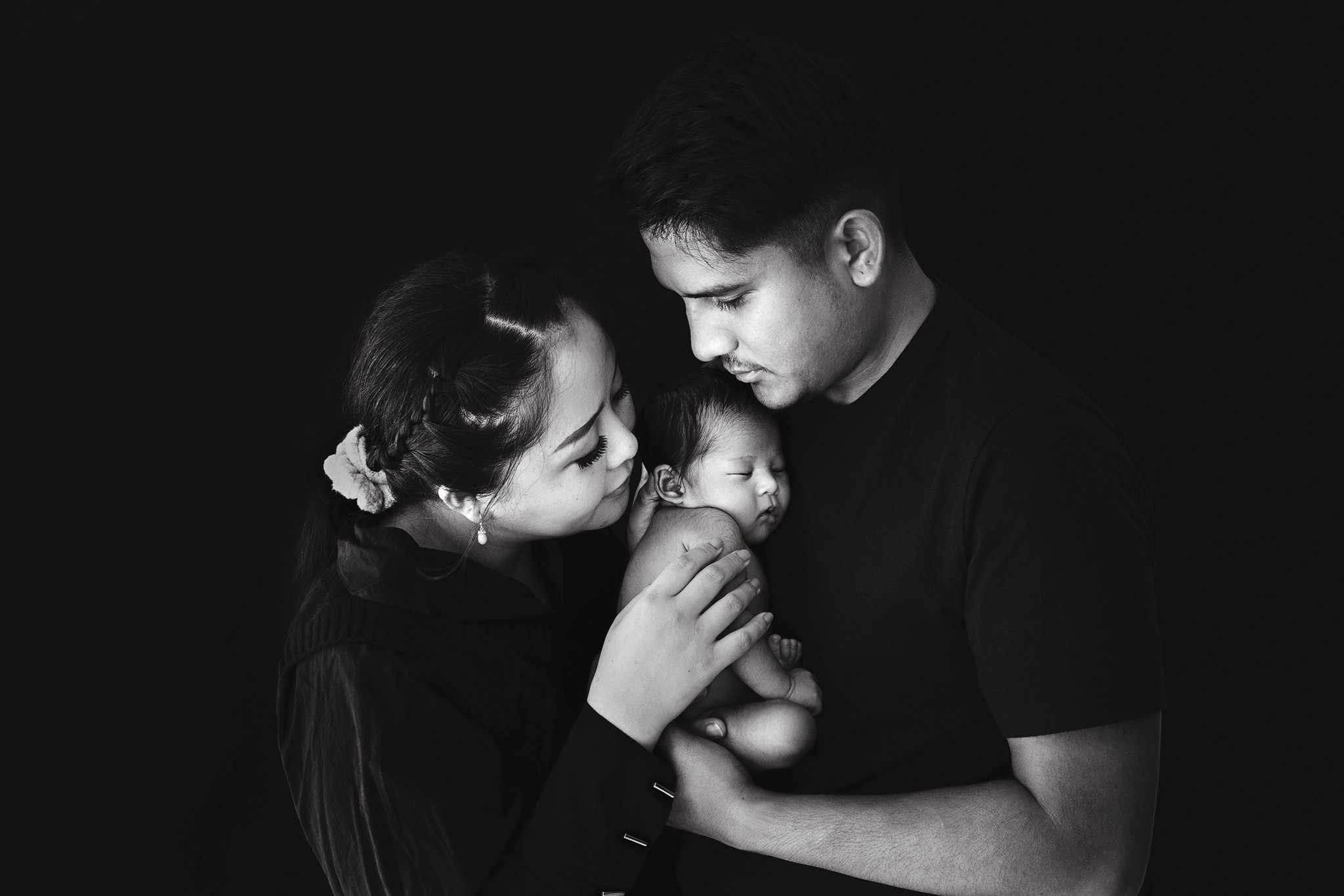 Black and white photo of a man and woman holding a newborn baby together affectionately, with dark background.