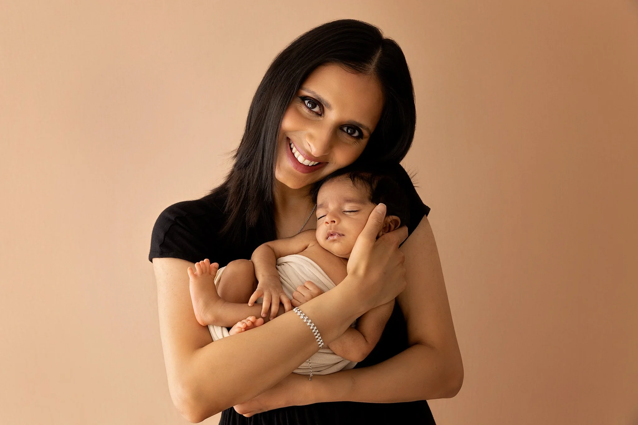 A woman with black hair and a black shirt holding a sleeping baby with dark hair and light skin, smiling in front of a beige background.