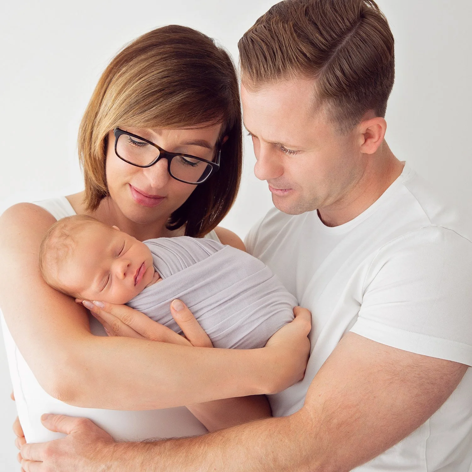 A mother holds her baby while the father wraps his arms around her, both gazing lovingly at their child.