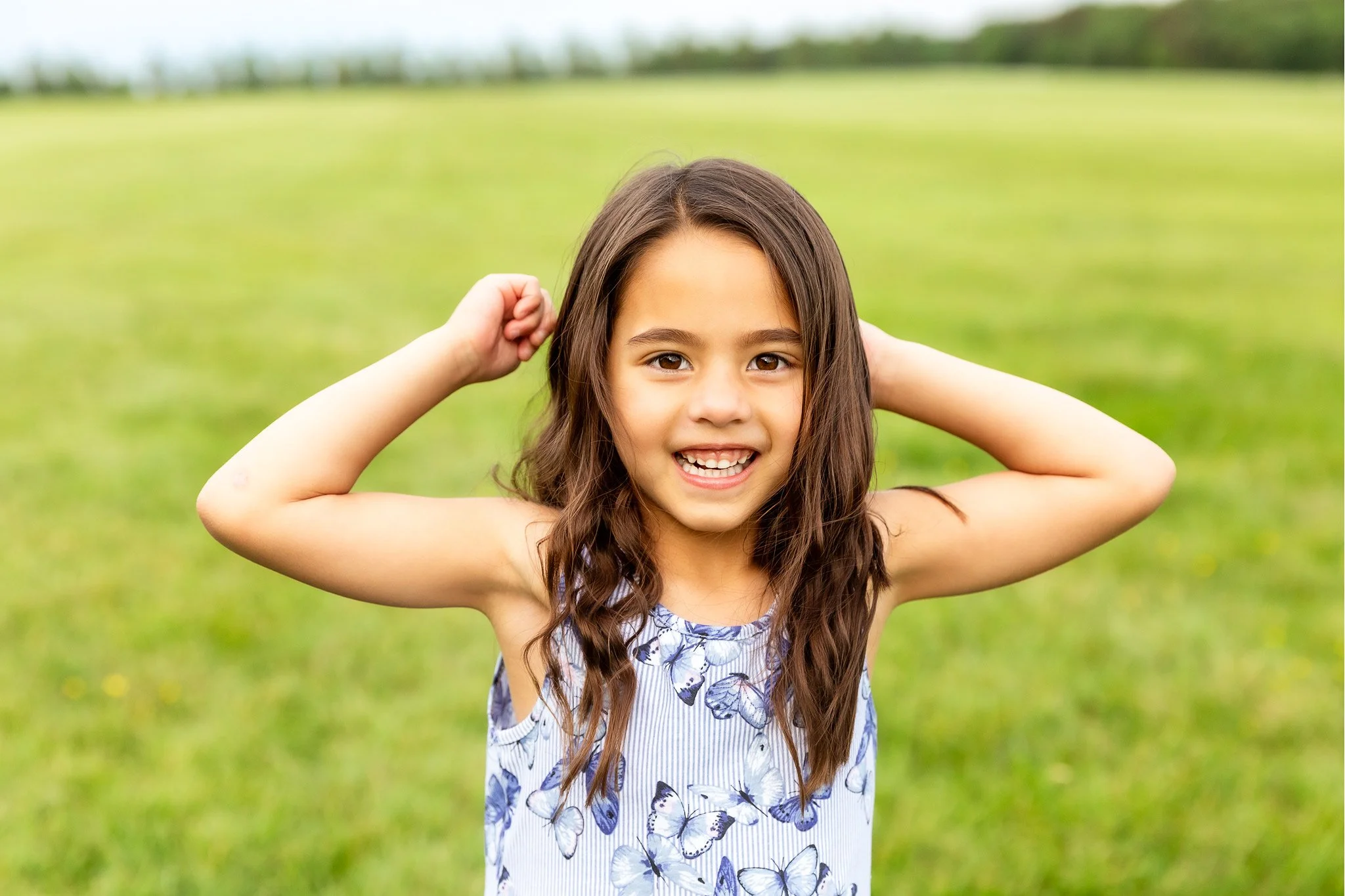 Young girl smiling outdoors in a green field, wearing a sleeveless dress with butterfly patterns, holding her hands behind her head.