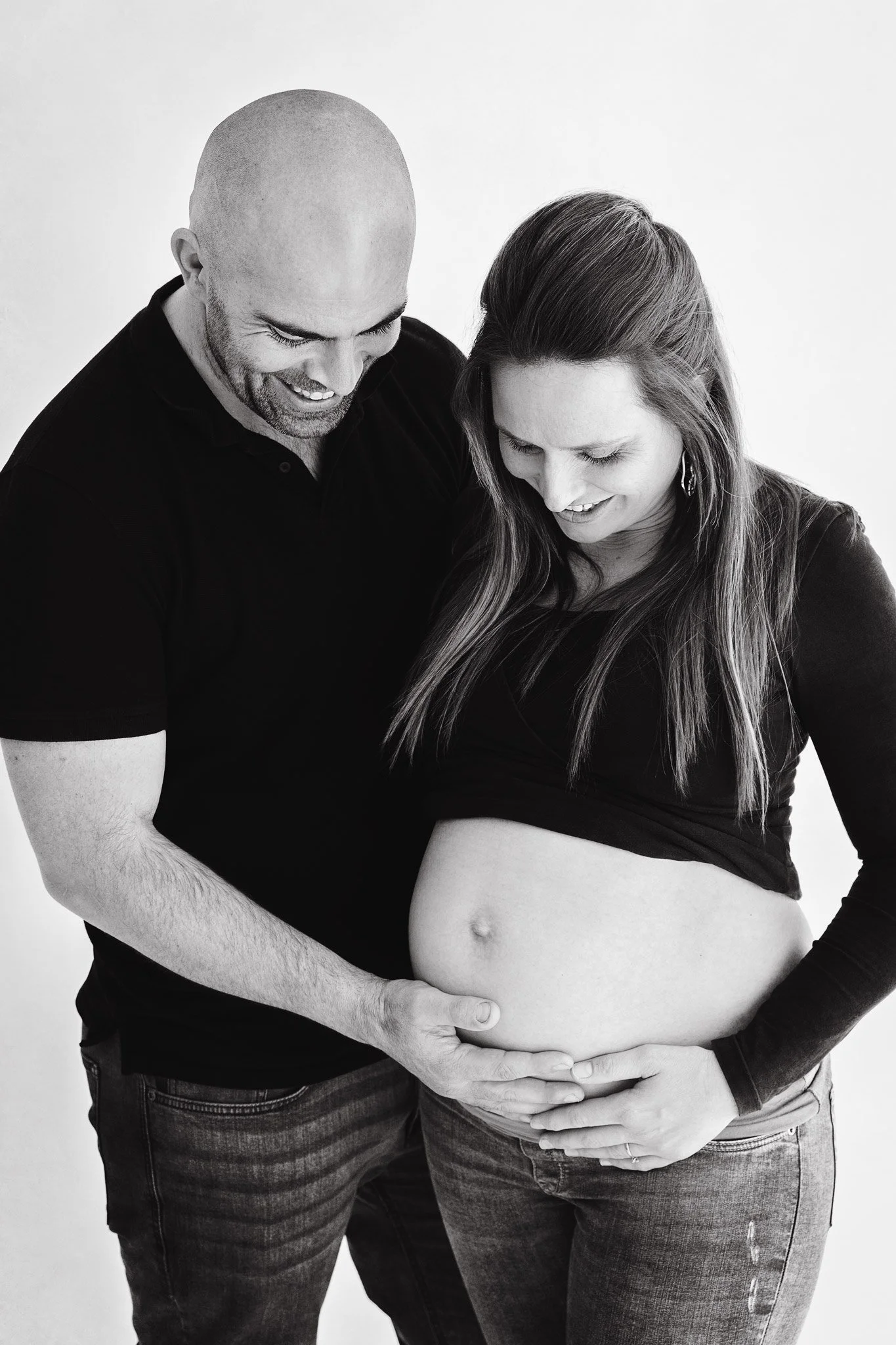 A smiling pregnant woman and a man touching her belly, both looking down at it, in black and white.