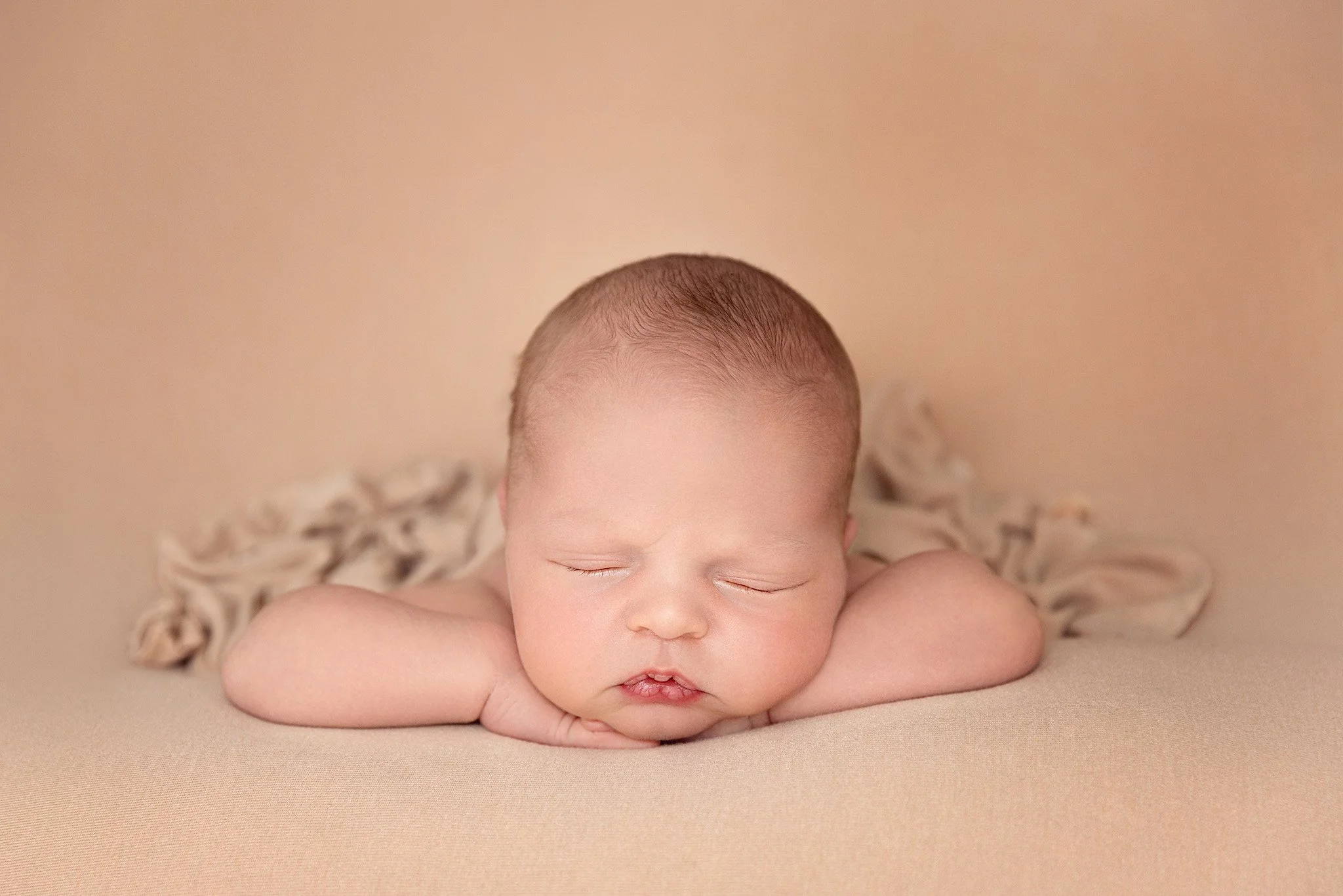A sleeping newborn baby with closed eyes resting on a beige surface with a matching beige background, arms under the chin, and a small amount of hair on the head.