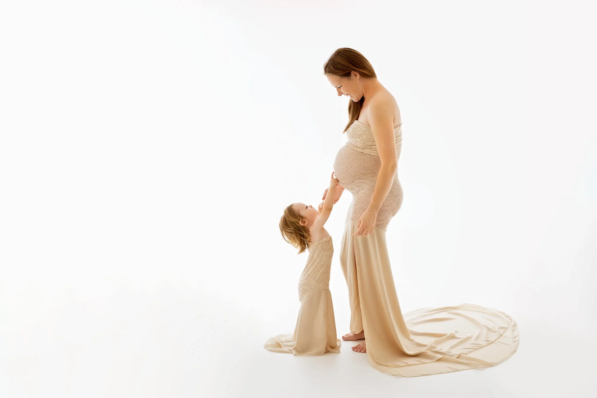 A pregnant woman and a small girl, both dressed in beige, are standing on a white background. The girl is touching the woman's pregnant belly, and they are looking at each other affectionately.