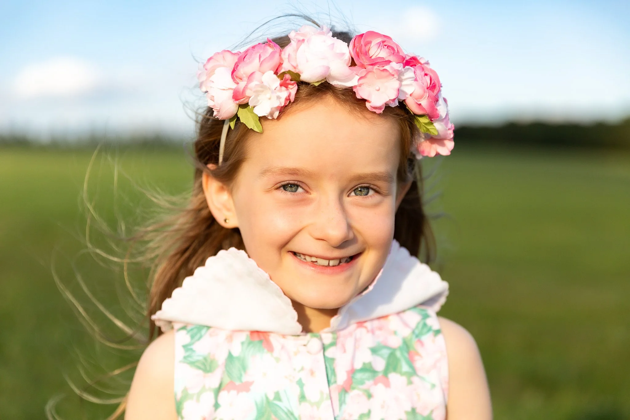 A girl with blue eyes smiling, wearing a pink and white flower crown, and a colorful sleeveless dress outdoors with grassy field and blue sky in the background.