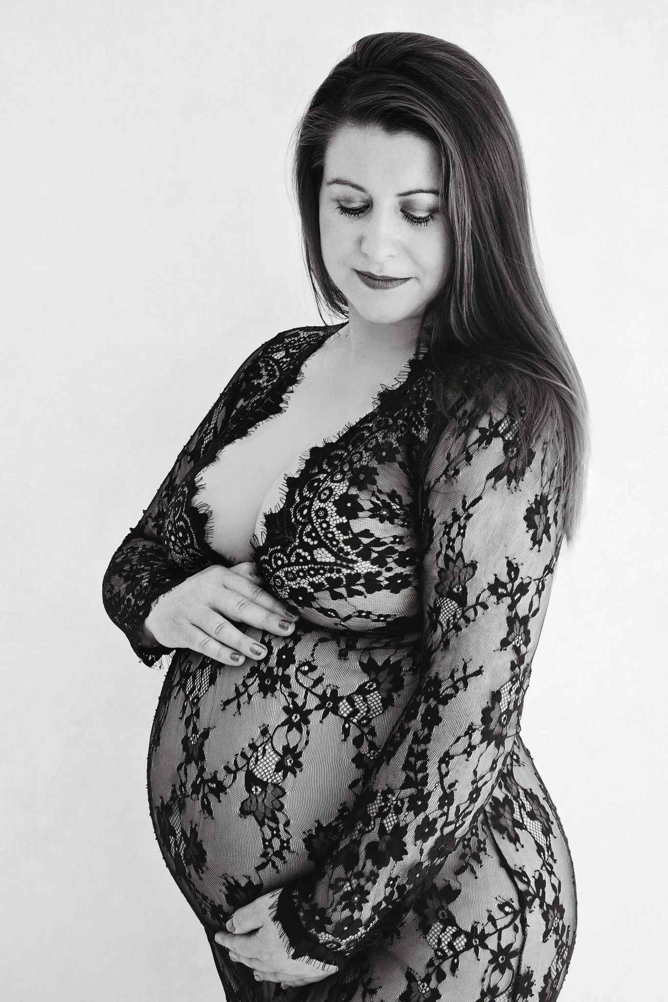 A pregnant woman in a black lace dress looking down and smiling, with her hand on her belly.