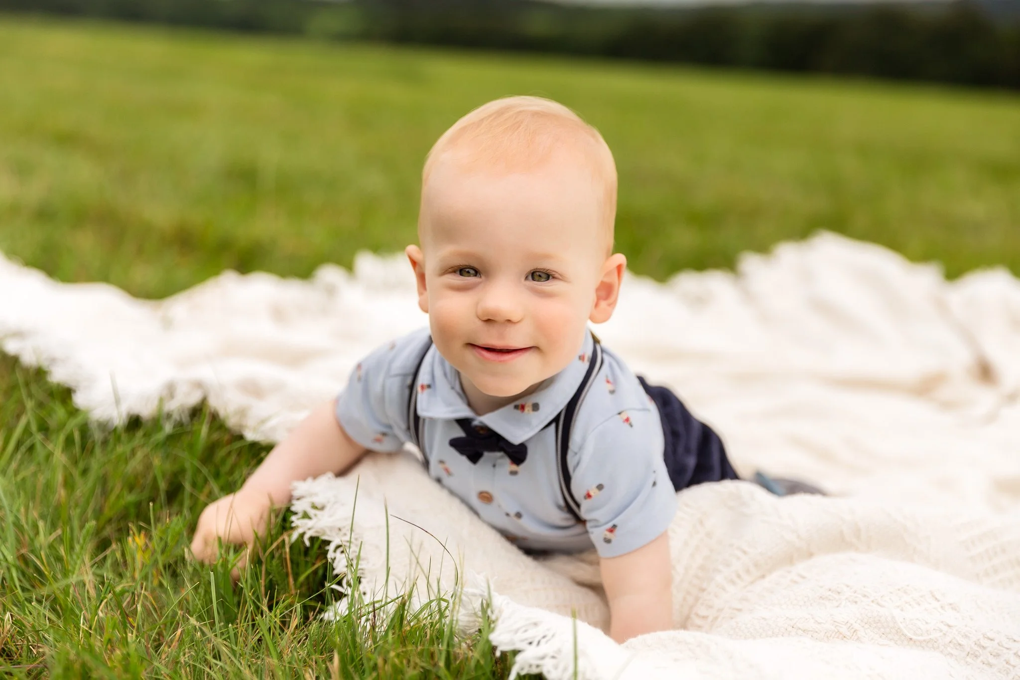 A young baby with blond hair and blue eyes lying on a white blanket on grass, smiling at the camera.
