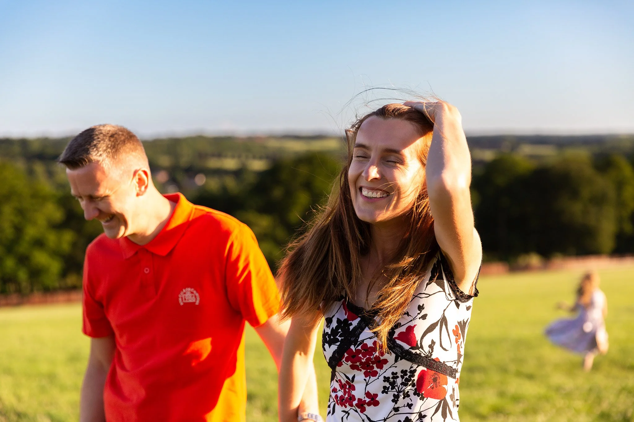 A smiling woman with long brown hair in a floral dress, holding her hair back with one hand, is outdoors during daylight. Next to her is a man in a bright orange shirt, laughing. A young girl in a white dress is in the background running on a grassy 