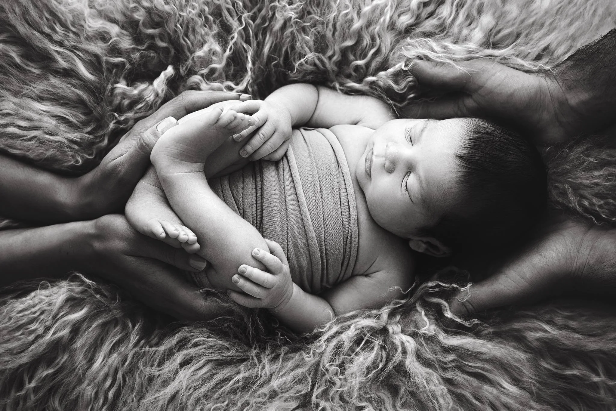 A baby lying on a soft, fluffy blanket with eyes closed, being gently held by an adult's hands, in black and white.