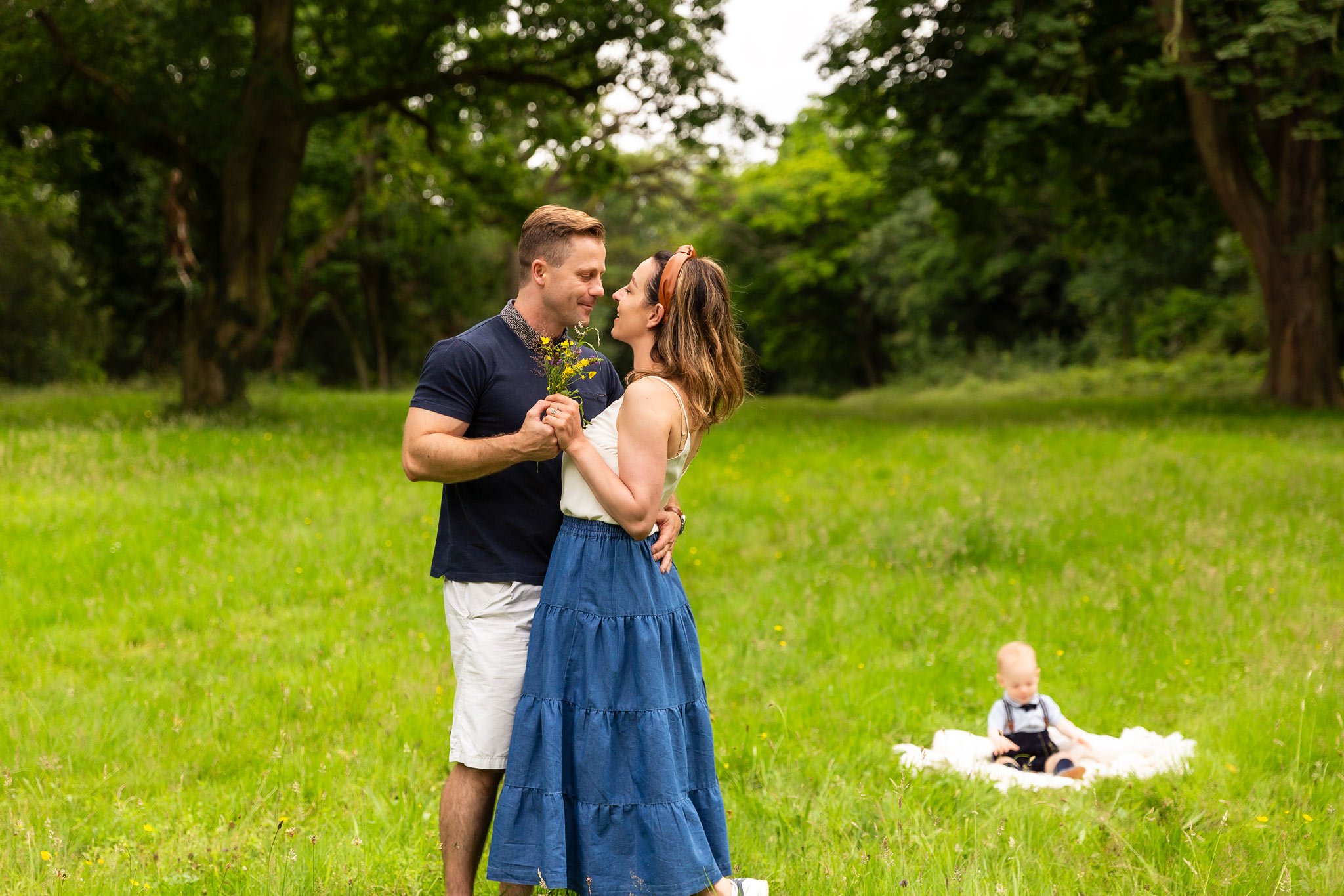 A couple holding flowers dancing in a green park with a baby sitting on a fluffy white blanket in the background.