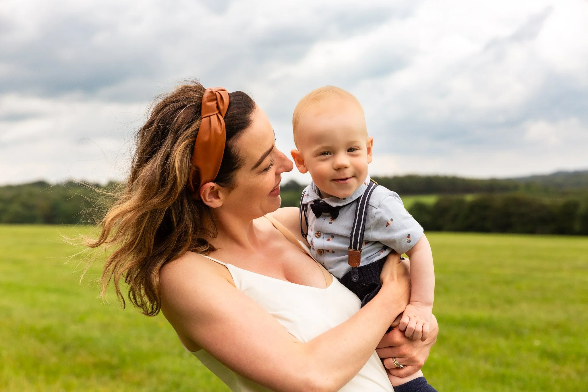 A smiling woman with brown hair wearing a brown headband holding a young boy dressed in a light blue shirt with suspenders and a bowtie in a green field with cloudy sky in the background.