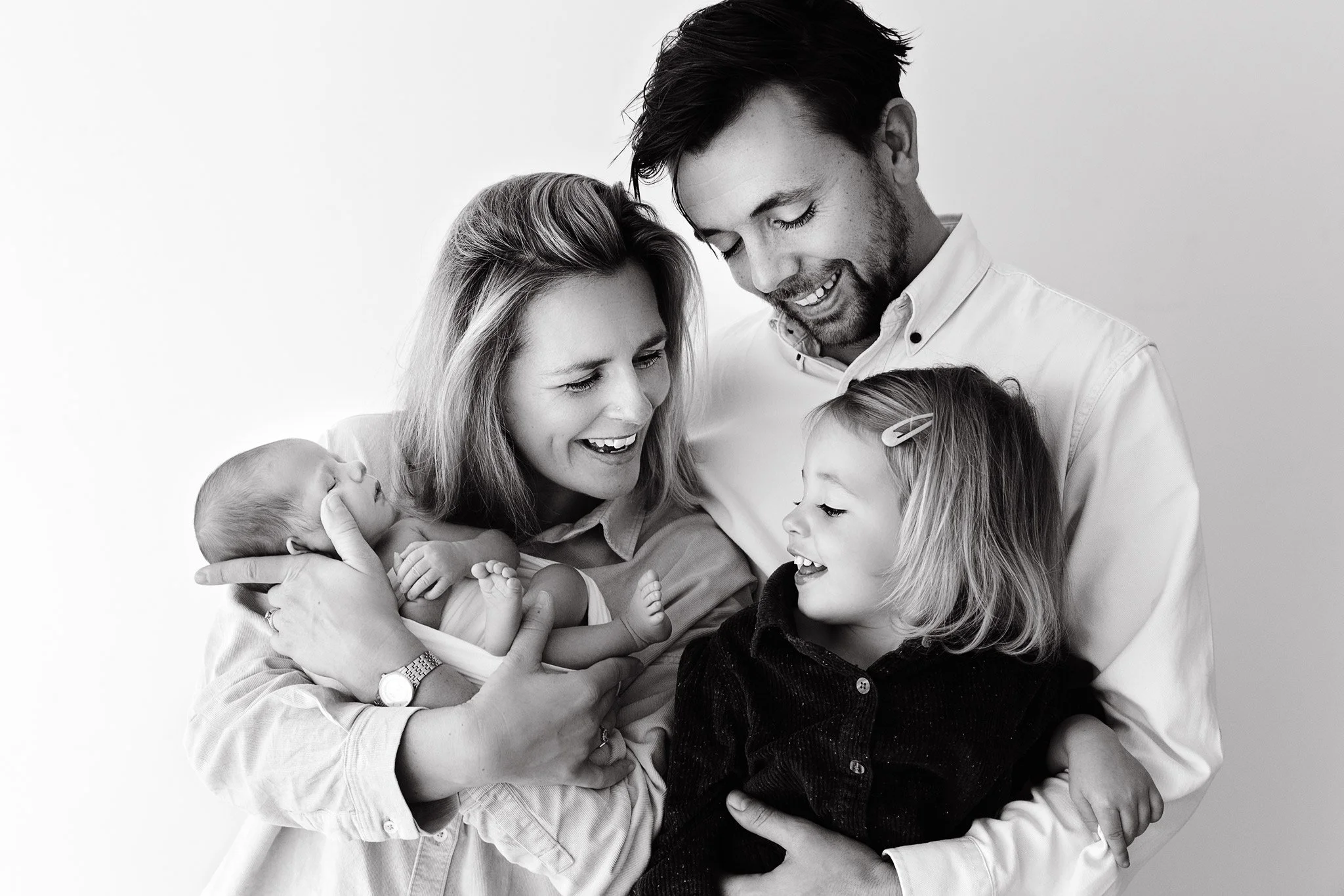 A family of four, including a woman, a man, a young girl, and a newborn baby, smiling and looking at each other in a close, joyful pose.