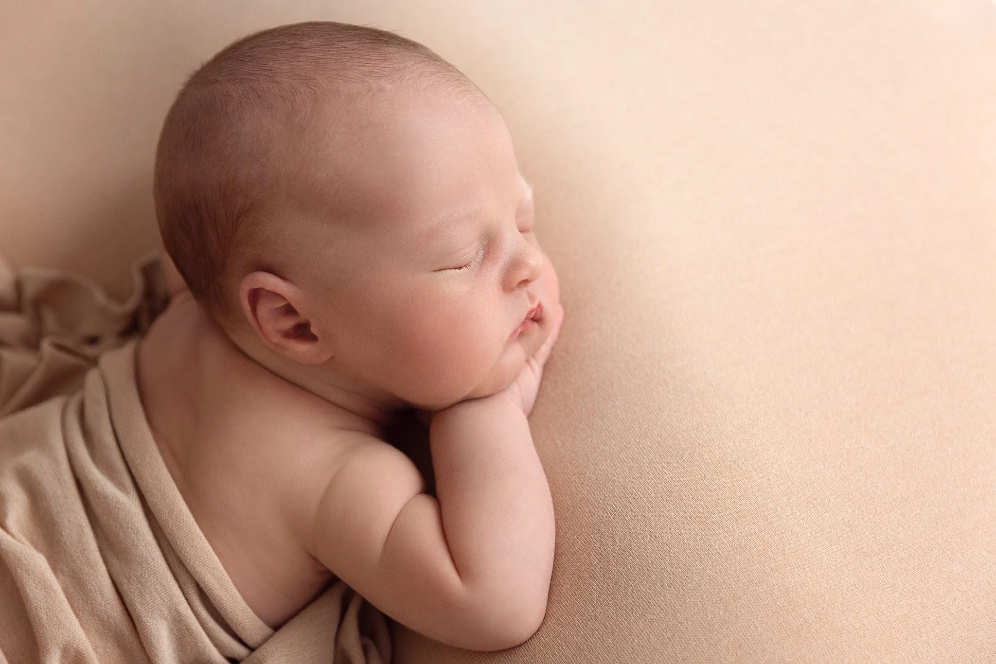 A sleeping baby with a hand under their chin, lying on a soft peach-colored surface, wearing a beige outfit.