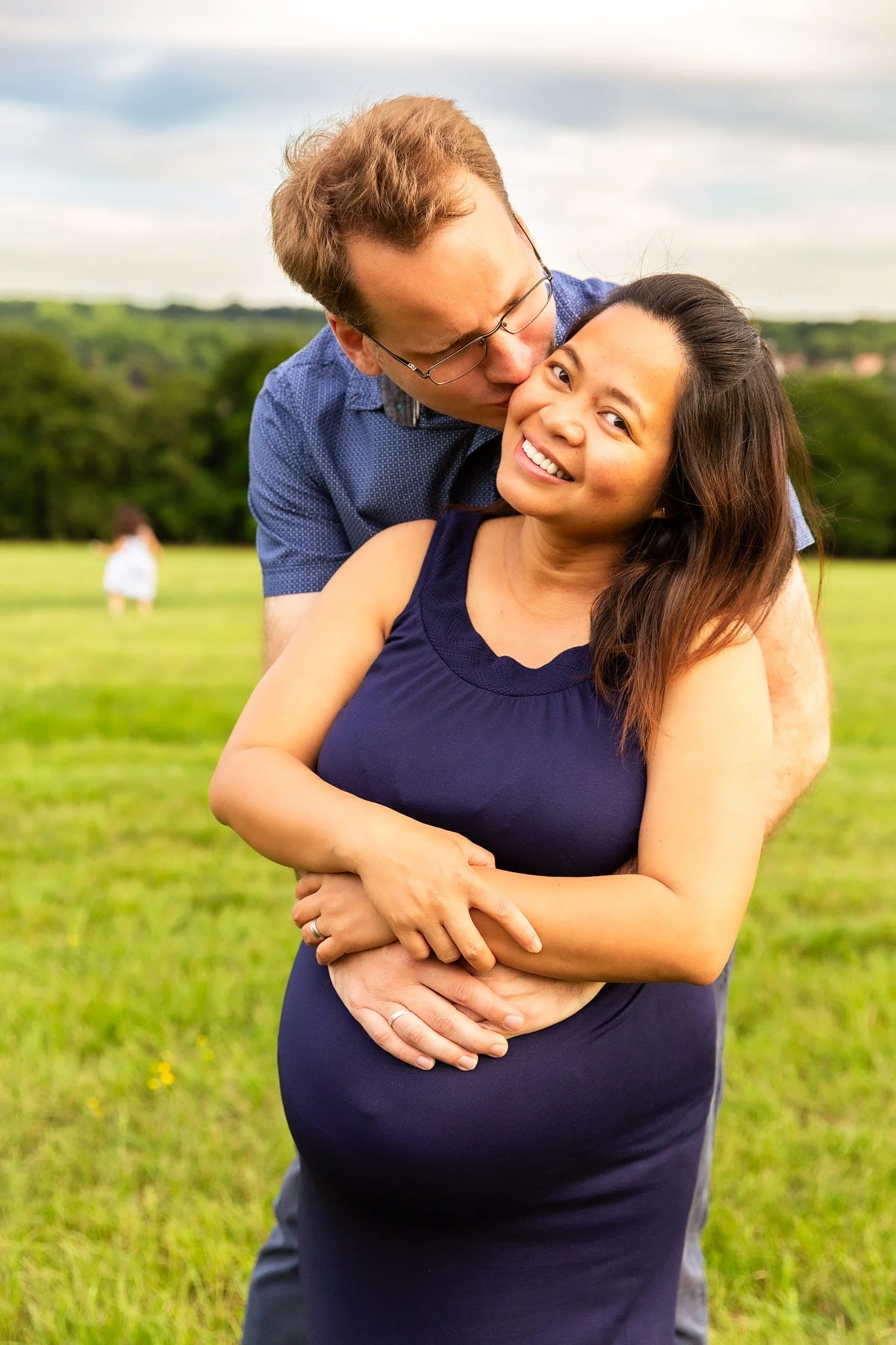 A happy couple in a green field, the man kissing the woman's forehead, she is smiling, and she appears to be pregnant.
