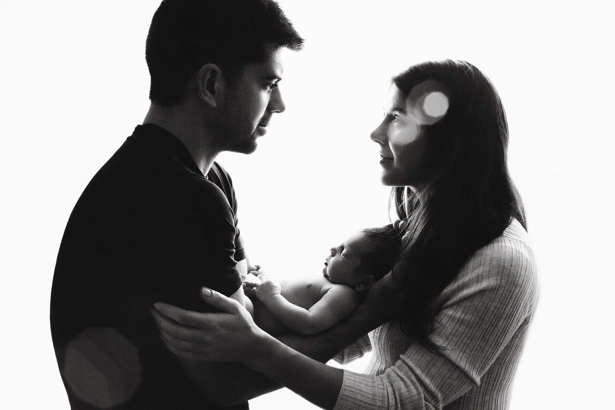 Silhouette of a family with a father, mother, and baby facing each other, with the mother holding the baby, against a bright background.