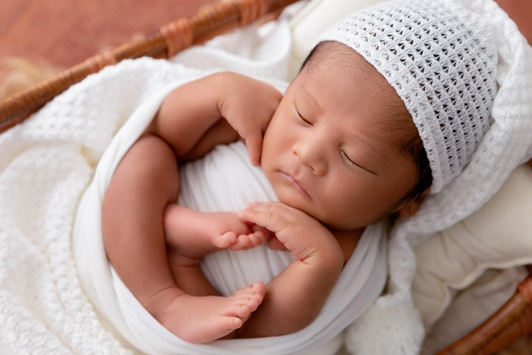 A sleeping infant with Asian features, wearing a white hat and shirt, lying on a soft blanket inside a wicker basket.