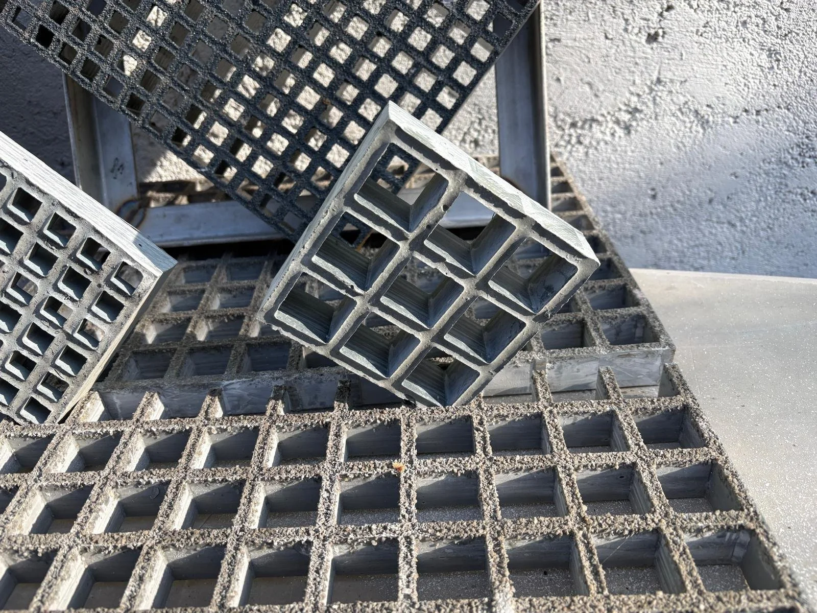 Riempimento di mattonelle di plastica grigia con fori quadrati, disposte su una superficie di cemento, con alcune mattonelle impilate su altre.