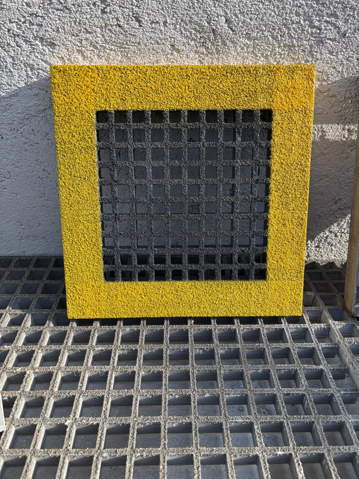 Una grata di metallo con una cornice gialla, posta su una superficie di griglia metallica, davanti a un muro bianco.