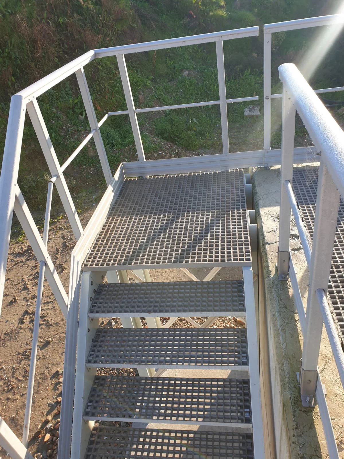 Scala metallica esterna con ringhiera e parapetto, vista dall'alto, con terreno e vegetazione sullo sfondo.