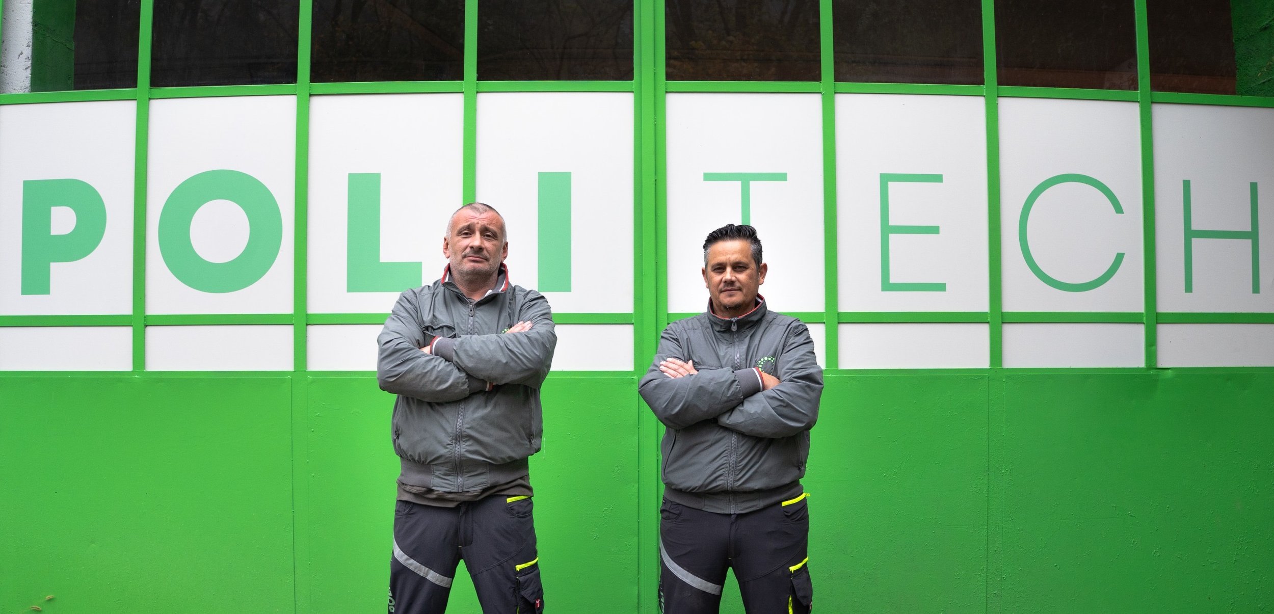 Due uomini in abbigliamento nero e grigio con braccia incrociate davanti a un muro verde con lettere bianche che formano la parola "POLITECH".