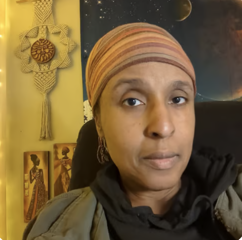 A Black woman looking directly at camera in a green and black sweatshirt and red and orange headwrap.