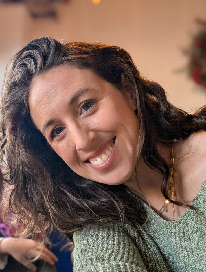 A white woman with long brown, wavy hair tilting to the left in frame and smiling directly at the camera.