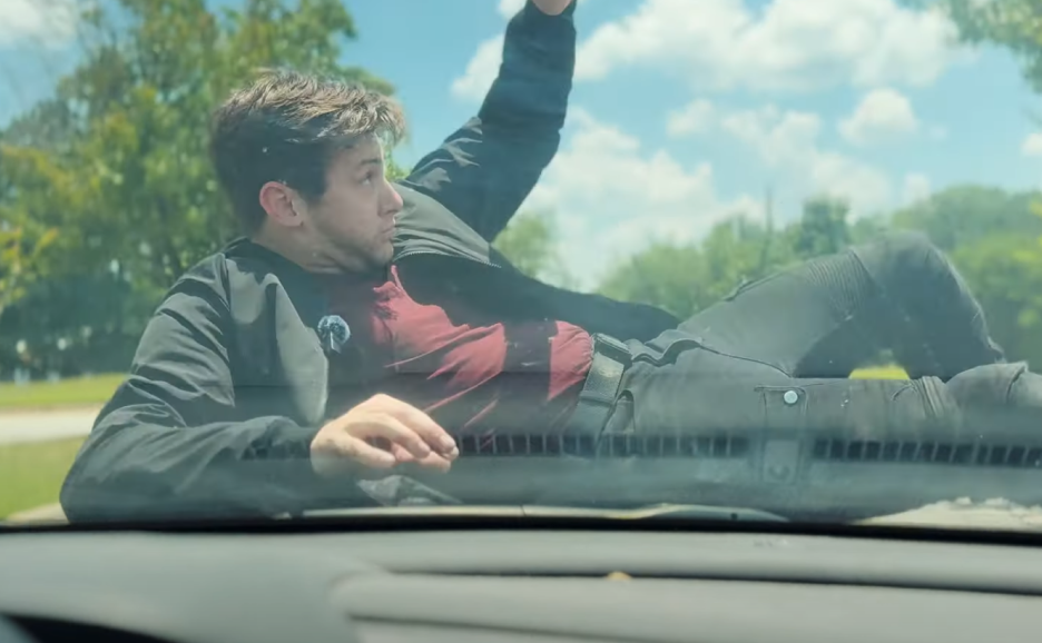 Man lying on the hood of a car, outdoors in a park, reaching across the windshield toward the sky with trees and a cloudy sky in the background.